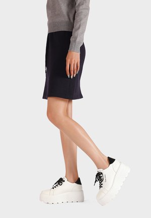 Persona che indossa una gonna nera, un maglione grigio a maniche lunghe e sneakers bianche platform con lacci neri, in piedi su una gamba.