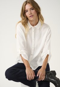 White, textured button-up shirt with a collar and roll-up sleeves, paired with dark denim pants and black ankle boots.
