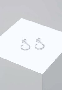Petites boucles d'oreilles créoles en argent avec attaches papillon, présentées sur un cube blanc sur un fond gris clair.
