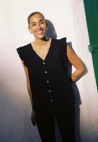 Personne souriante aux cheveux courts et tatouages sur les bras, portant un haut sans manches noir boutonné, debout contre un mur blanc au soleil.