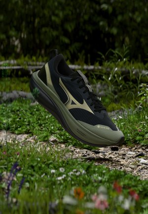 Zapatilla de correr negra y verde oliva con etiqueta Gore-Tex flotando sobre un camino rocoso rodeado de plantas y flores verdes.