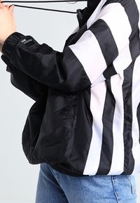 Svart- och vitrandig vindjacka med dragkedja fram, snörning i huvan och elastiska ärmmuddar. Tillverkad av lätt, glänsande tyg.