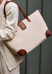 Beige tote taske med brune læderdetaljer, herunder håndtag og nederste hjørner. Har en knaplukning med et logo. 