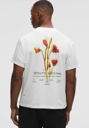 T-shirt en coton blanc avec un motif floral en rouge, jaune et vert au dos, comportant le texte "détours bienvenus" et le logo de la marque.