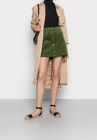 Grön kordflodermini kjol med framknappar, i kombination med en svart polotröja och en beige trenchcoat. Svarta espadrillosandaler fulländar looken.