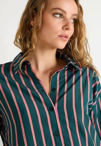 Camisa a rayas verdes con rayas verticales en rosa y blanco, un cuello clásico y botones negros en el frente. Textura suave.