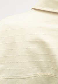 Tissu couleur crème avec de subtiles rayures horizontales, présentant une surface texturée et un design à col. Des coutures apparentes ajoutent du détail.