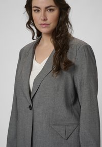 Blazer gri cu dungi subțiri, cu rever clasic, închidere cu un singur nasture și un buzunar frontal cu detaliu triangular. Material ușor, croială ajustată.