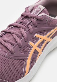 Baskets de sport violettes avec une tige en mesh ornée d'accents orange, des lacets texturés et une semelle en caoutchouc blanche contrastante.