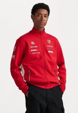 Red zip-up jacket with a high collar, featuring white side panels, multiple branded patches, and zippered pockets. Made of soft fabric.