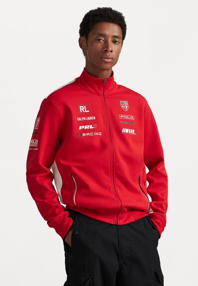 Rød zip-up jakke med højt krave, med hvide sidelapper, flere mærkede patches og lynlåsede lommer. Fremstillet af blødt stof.
