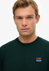 Camiseta de algodón verde oscuro con cuello redondo, mangas cortas y un colorido parche de logo bordado en el lado izquierdo del pecho.