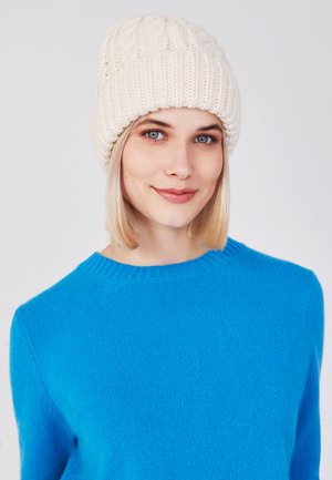 Off-white gebreide beanie met een kabelpatroon. Draagt een levendige blauwe trui met een ronde halslijn en lange mouwen.