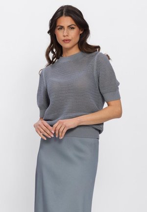 Femme portant un pull en tricot gris à manches courtes et une jupe gris assortie, debout les mains croisées devant elle.