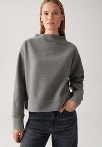 Giovane donna che indossa un ampio maglione grigio a maniche lunghe con polsini larghi e jeans neri, in piedi con una mano in tasca su uno sfondo semplice.