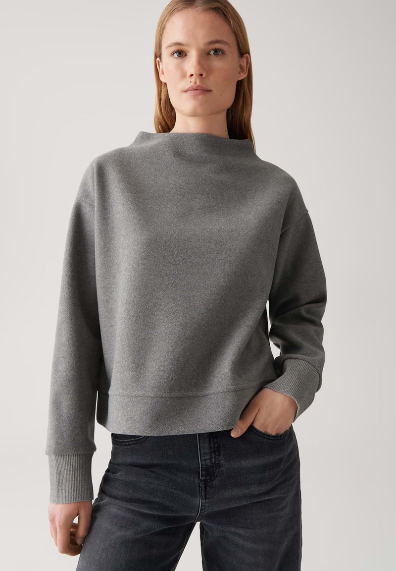 Giovane donna che indossa un ampio maglione grigio a maniche lunghe con polsini larghi e jeans neri, in piedi con una mano in tasca su uno sfondo semplice.