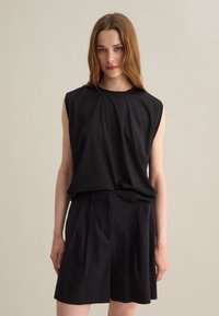 Top senza maniche nero realizzato in tessuto morbido, abbinato a pantaloni pantaloni neri su misura con pieghe e una vestibilità rilassata. Sfondo neutro.