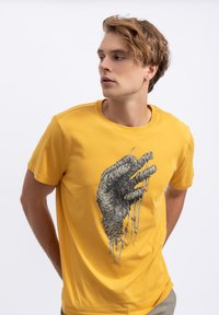 Geel katoenen t-shirt met een gedetailleerde zwarte grafiek van een hand met stringachtige elementen. Korte mouwen en een ronde halslijn.