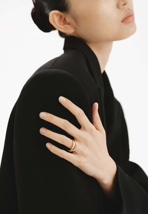 Anillo de oro con múltiples bandas, con acentos texturizados, llevado en una mano elegantemente exhibida que reposa sobre una prenda negra.