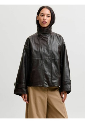 Femme portant une veste en cuir noire avec un col montant et des manches larges, associée à un pantalon beige ample, debout devant un fond uni.