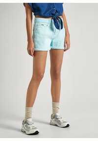 Pantalones cortos de mezclilla azul claro con vueltas en los puños, con cierre de botón y bolsillos laterales, combinados con calcetines beige y zapatillas deportivas grises gruesas.