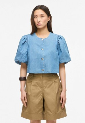 Kobieta ubrana w jasnoniebieską bluzkę na guziki z bufkami krótkich rękawów oraz beżowe szorty z wysokim stanem długości do kolan, stojąca na białym tle.