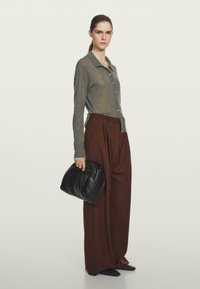 Graues langärmliges Hemd mit Knöpfen, braune hoch taillierte weite Hose, hält eine schwarze Clutch. Glatte Stofftextur, minimalistisches Design.