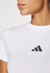 Bjela pamučna majica s okruglim izrezom i crnim Adidas logotipom, s glatkom teksturom i kratkim rukavima.
