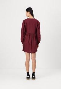 Vestido bordeaux com corpete justo e saia franzida, apresentando mangas bufantes e decote redondo. Estilizado com meias pretas e ténis.