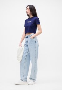 Tamsiai mėlynas trumpas marškinėliai su tekstu, šviesiai mėlynos aukšto liemens džinsai, balti sportbačiai ir grietinėlės spalvos džemperis laikomas vienoje rankoje.