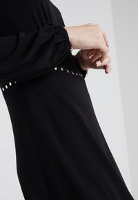 Robe noire avec de longues manches froncées et une taille ajustée. Accentuée de clous métalliques le long de la taille. Tissu lisse et mat.