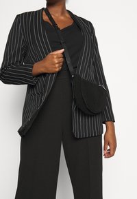 Černý pruhovaný blazer přes černý top, doplněný širokými kalhotami a malou černou crossbody taškou s oblým tvarem a zipem.