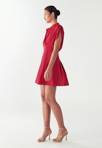 Rotes, kurzes Kleid mit kurzen Ärmeln, plissiertem Rock, Kragen und Knopfverschluss. Glattes Material und taillierte Passform. Getragen mit nude-farbenen Riemchen-Heels.