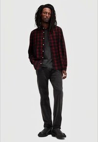 Camicia a quadri nera e rossa con bottoni, T-shirt grigia sotto, jeans neri e stivali neri. Il modello si trova su uno sfondo neutro.