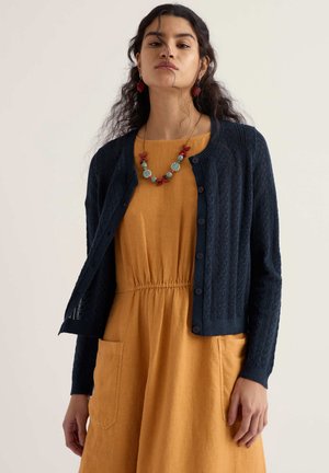 Moteris su tamsiais garbanotais plaukais vilki garstyčių spalvos suknelę su priekinėmis kišenėmis, tamsiai mėlyną megztinį su sagomis ir gėlių karoliukų vėrinį bei auskarus.