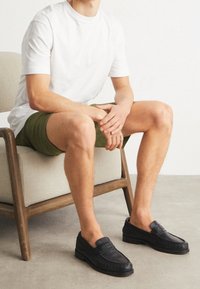 Czarne skórzane loafersy z detalami szycia, zaokrąglony czubek i niska podeszwa, noszone z zielonymi szortami, na jasnoszarym podłożu.