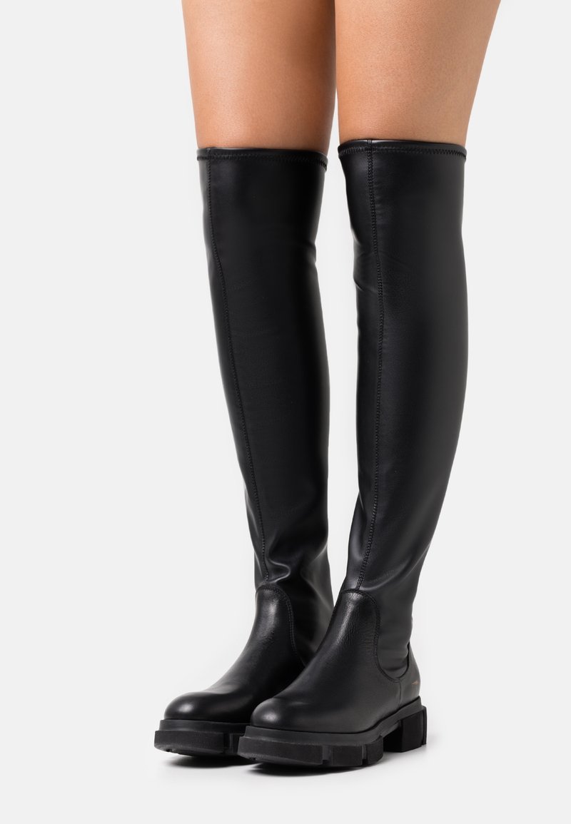 Bottes hautes en cuir noir avec semelles épaisses portées sur des jambes nues, qui montent au-dessus du genou sur un fond clair uni.