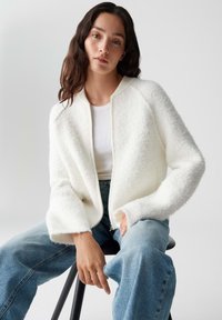 Witte, fluffy cardigan met een ritssluiting aan de voorkant, ribbelkraag en brede mouwen, gecombineerd met lichtblauwe hoge taille jeans.