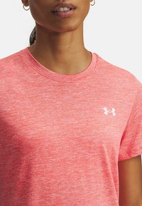Camiseta deportiva rosa hecha de tela transpirable, con cuello redondo, mangas cortas y un logo blanco de Under Armour en el frente.