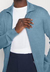 Cardigan con zip lavorato a maglia in azzurro, caratterizzato da un motivo testurizzato e polsini a coste. Indossato sopra una t-shirt bianca con pantaloni blu navy.