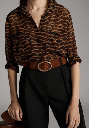 Femme portant un chemisier marron et noir à motifs, rentré dans un pantalon noir taille haute avec une ceinture marron ornée d'une boucle ovale dorée.