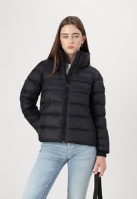 Svart boblejakke med horisontalt kviltedesign, høy krage, frontglidelås og glidelåslommer på sidene. Brukt med lyseblå jeans.