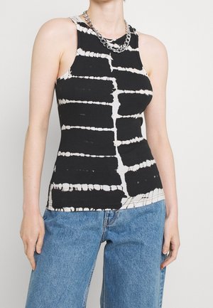 Femme portant un haut sans manches tie-dye noir et blanc avec un collier en chaîne argentée et un jean bleu, debout devant un fond uni.