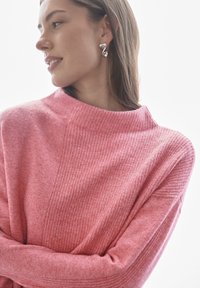 Lichtroze trui met een hoge hals en gestructureerde ribbels. Gladde stof met een subtiele glans. Model draagt zilveren spiraaloorbellen.