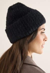 Musta neulottu myssy, jossa on pörröinen texture, renno kaava ja hienovarainen kimalle. Kantaa hieman vinoon neutraalia taustaa vasten.