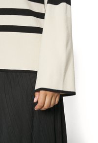 Main avec des ongles rose clair à côté d'une manche de pull côtelé noir et blanc et d'une jupe noire plissée.