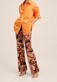 Chemise orange à boutons avec manches retroussées, associée à un pantalon évasé orné d'un motif floral cachemire coloré sur un fond sombre.