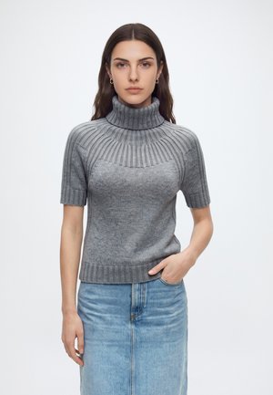 Jeune femme aux longs cheveux bruns porte un pull gris à col roulé à manches courtes et une jupe en jean bleu clair, debout avec une main dans la poche.