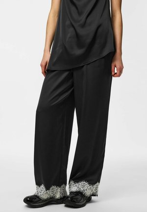 Femme portant un pantalon large en satin noir avec un bord en dentelle blanche à l'ourlet, assorti à un top en satin noir et des baskets noires.