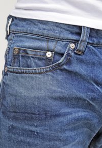 Jeans de mezclilla en azul medio con una textura desgastada, que cuentan con herrajes plateados, un pequeño bolsillo frontal y detalles de costura en contraste.